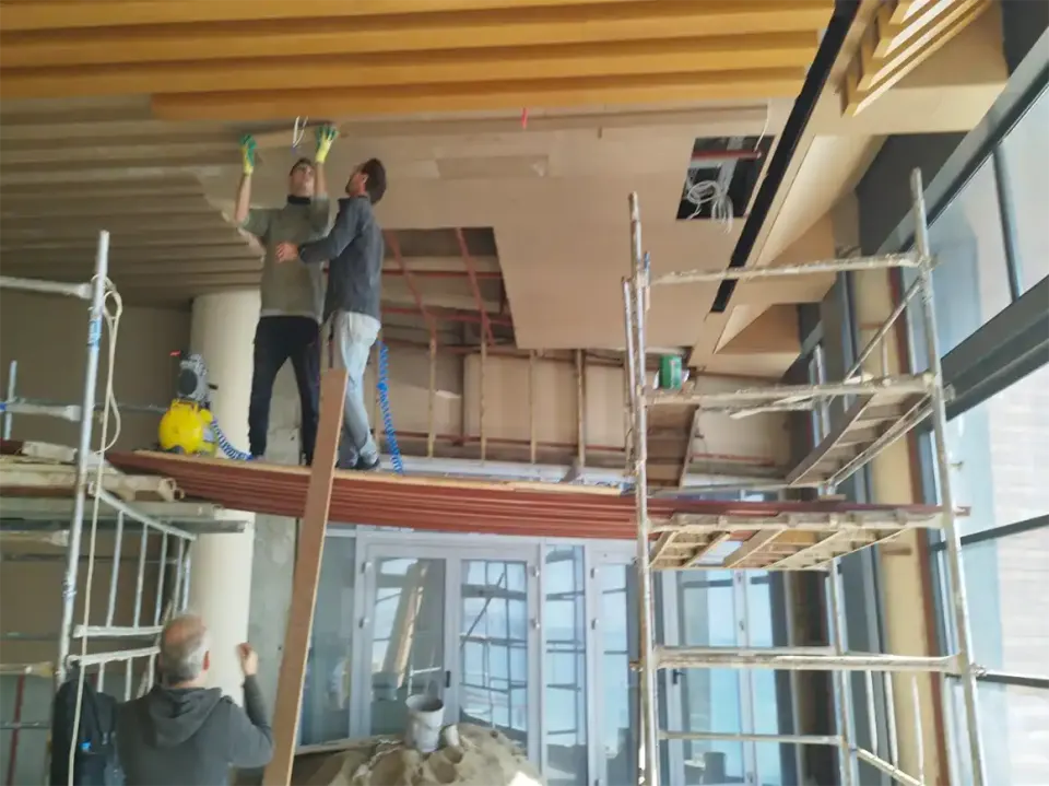 Two workers on a scaffold fixing wooden cladding boards to the ceiling by a glass facade